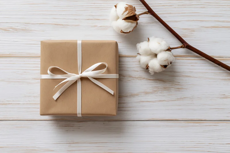 Kraft paper gift box tied with white satin ribbon on whitewashed wood surface with cotton plant branch