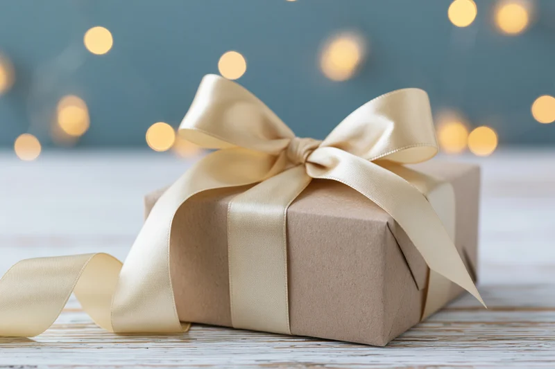 Kraft paper gift box with champagne satin ribbon bow on wooden surface against blurred warm lights