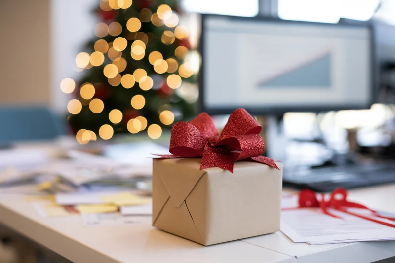 Gift box with red glitter bow on office desk near christmas tree and computer