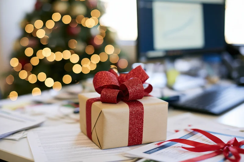 Gift box with red glitter bow on office desk near computer and christmas tree