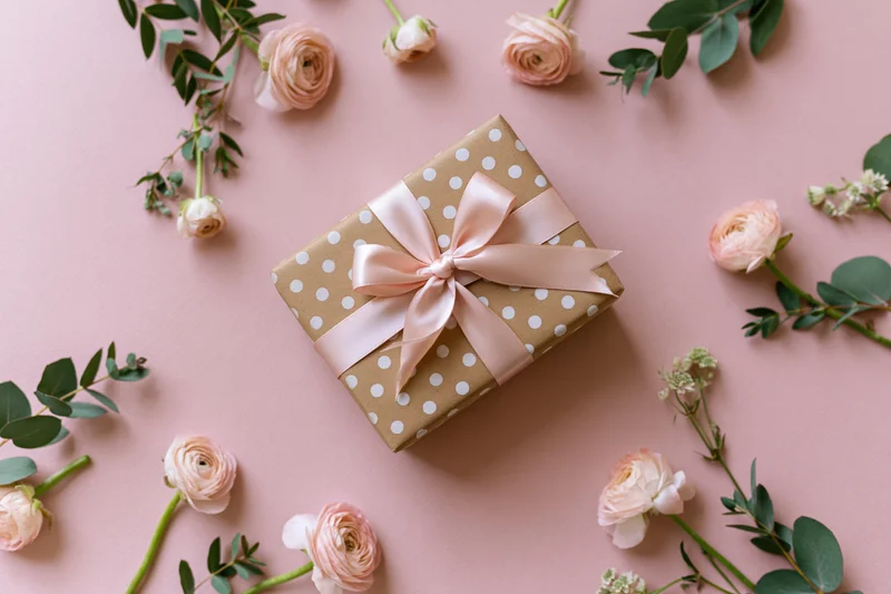 Gift box wrapped in kraft paper with white polka dots and pink ribbon bow, surrounded by roses and eucalyptus on pink surface