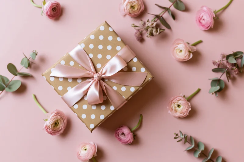 Gift box wrapped in kraft paper with white polka dots and pink ribbon bow, surrounded by pink roses and eucalyptus on a soft pink surface