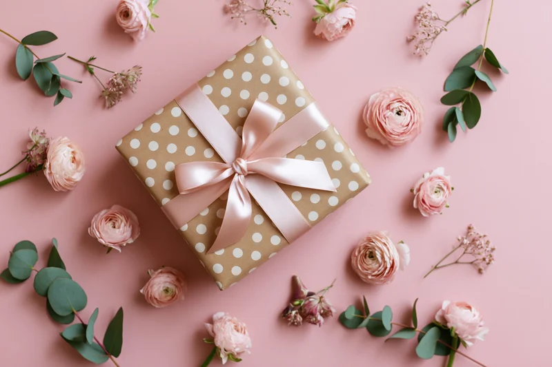 Gift box wrapped in kraft paper with white polka dots and pink ribbon bow, surrounded by pink roses and eucalyptus on a pink background