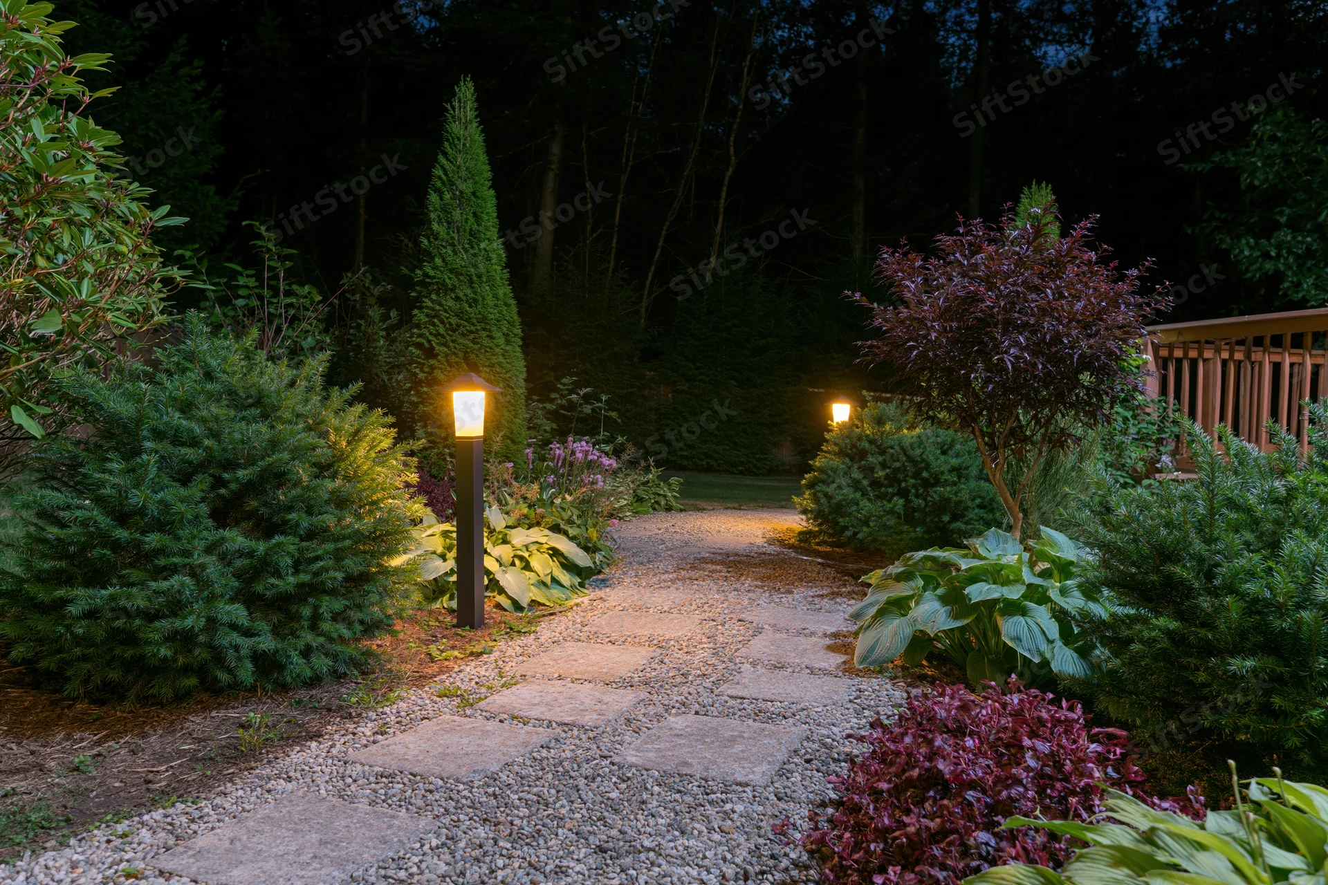 Stone and gravel garden path at night illuminated by tall bollard lights among shrubs and trees