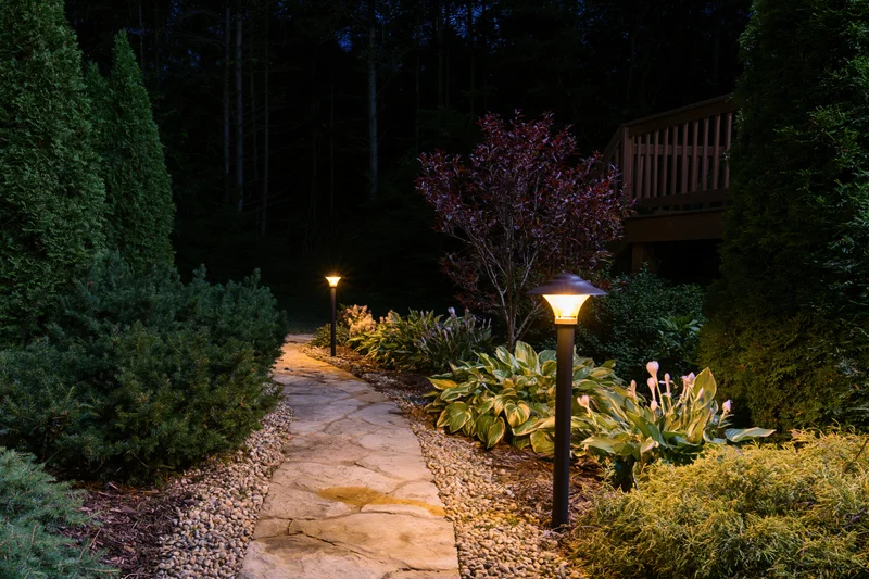 Stone garden path illuminated by tall bollard lamps at night surrounded by shrubs and plants