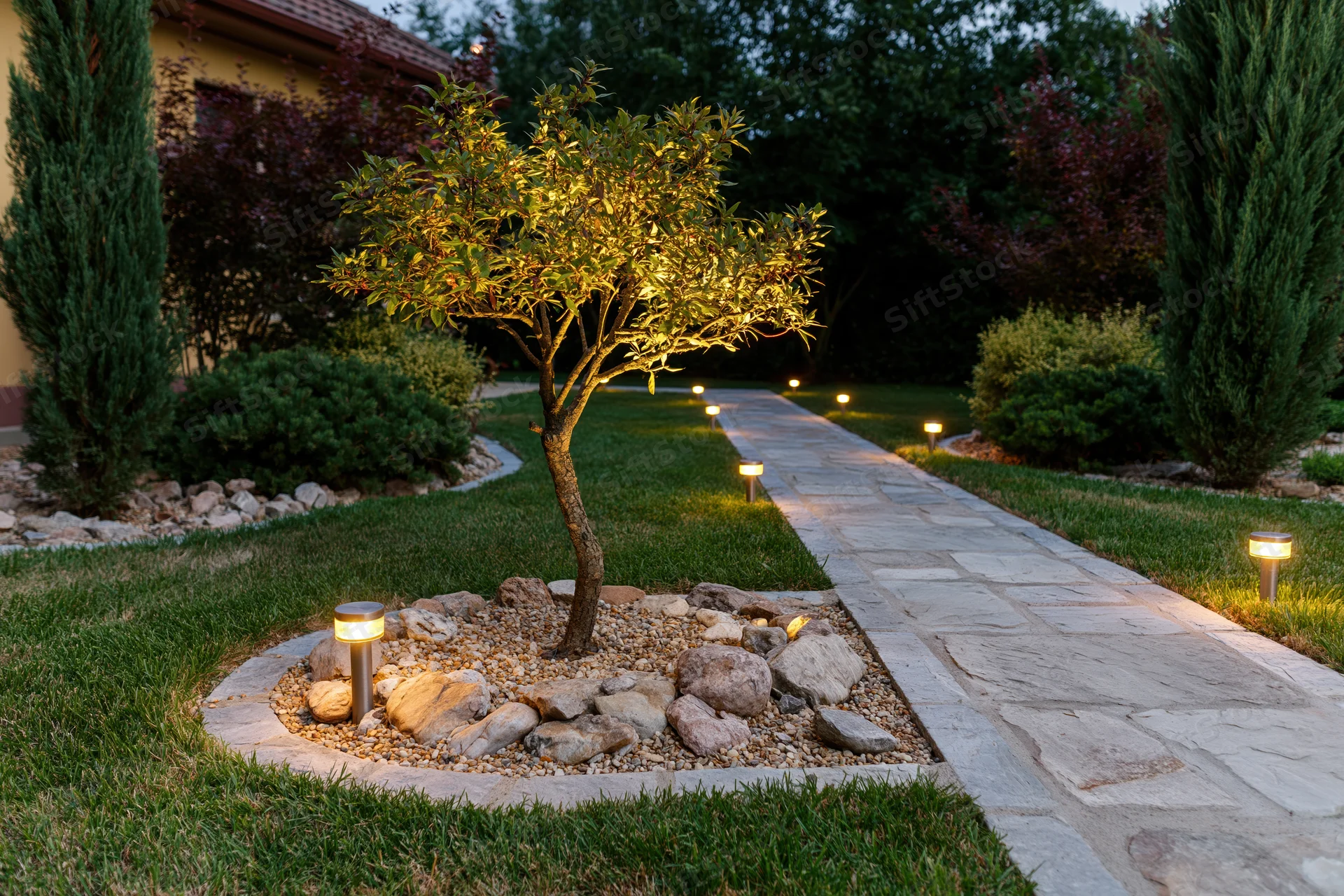 Illuminated stone pathway through a landscaped garden at night with bollard lights and a small tree in a rock bed