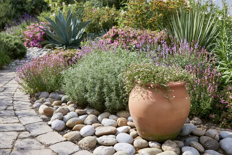 Large terracotta urn with trailing flowers in a garden bordered by stones and greenery along a stone pathway