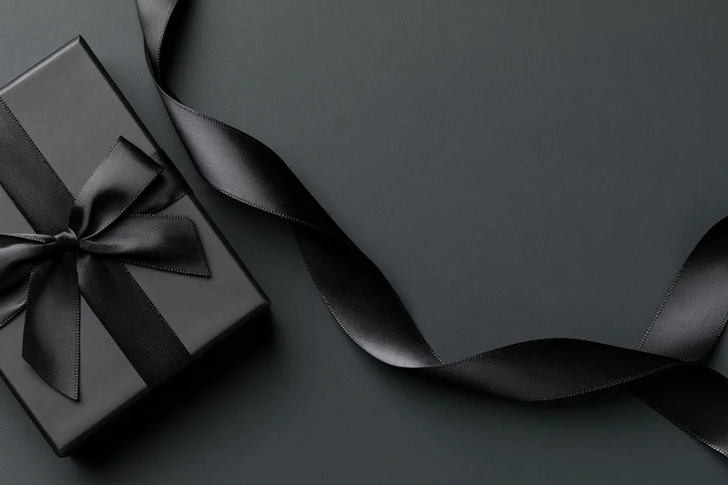 Matte black gift box with satin ribbon bow on dark surface, flat lay view