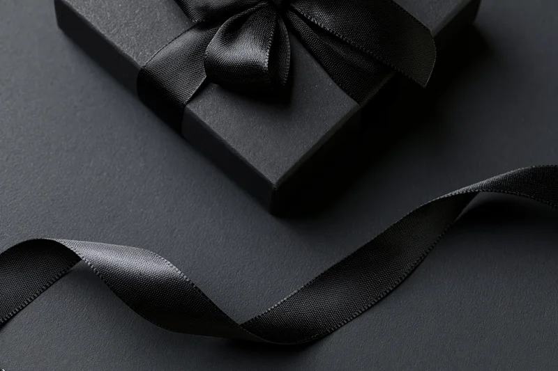 Matte black gift box with satin ribbon bow on dark surface, flat lay view