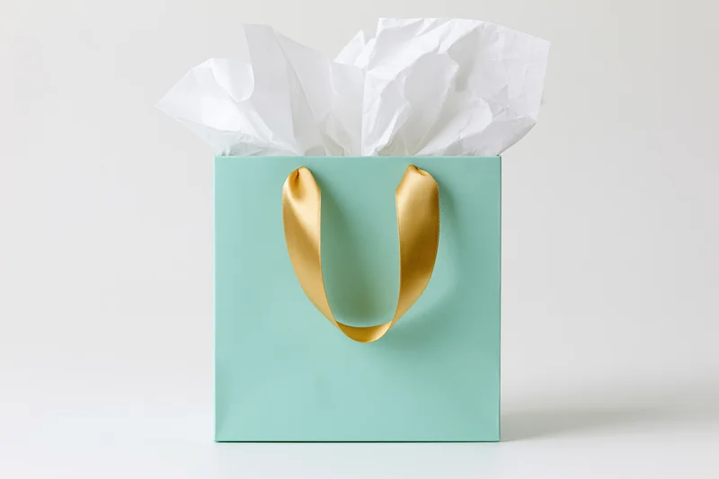 Mint green gift bag with gold satin ribbon handles and white tissue paper on white background