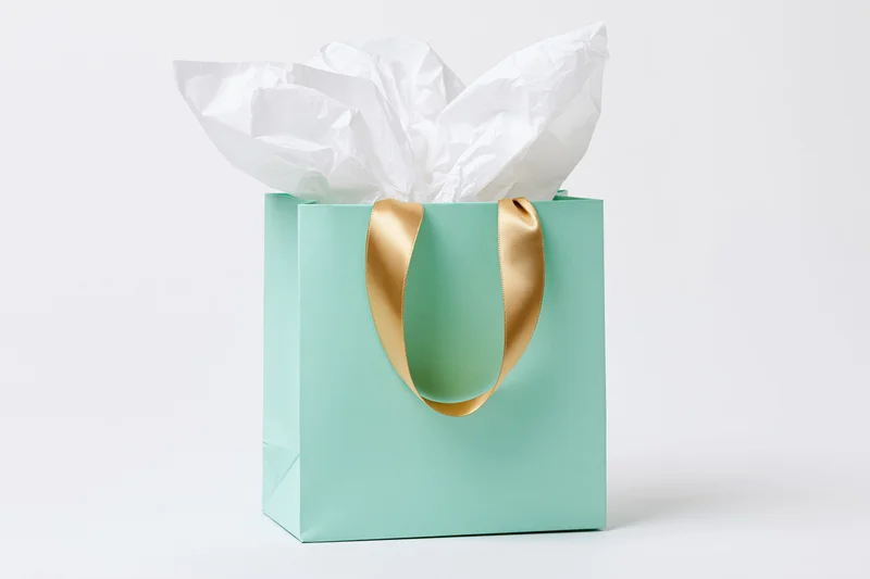 Mint green gift bag with gold satin ribbon handles and white tissue paper on white background