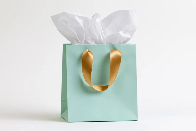 Mint green gift bag with gold satin ribbon handles and white tissue paper on white background