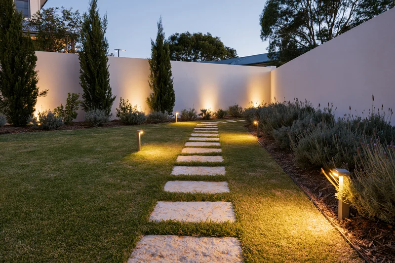 Modern residential garden at dusk with stepping stones pathway through lawn and illuminated shrubs along white wall