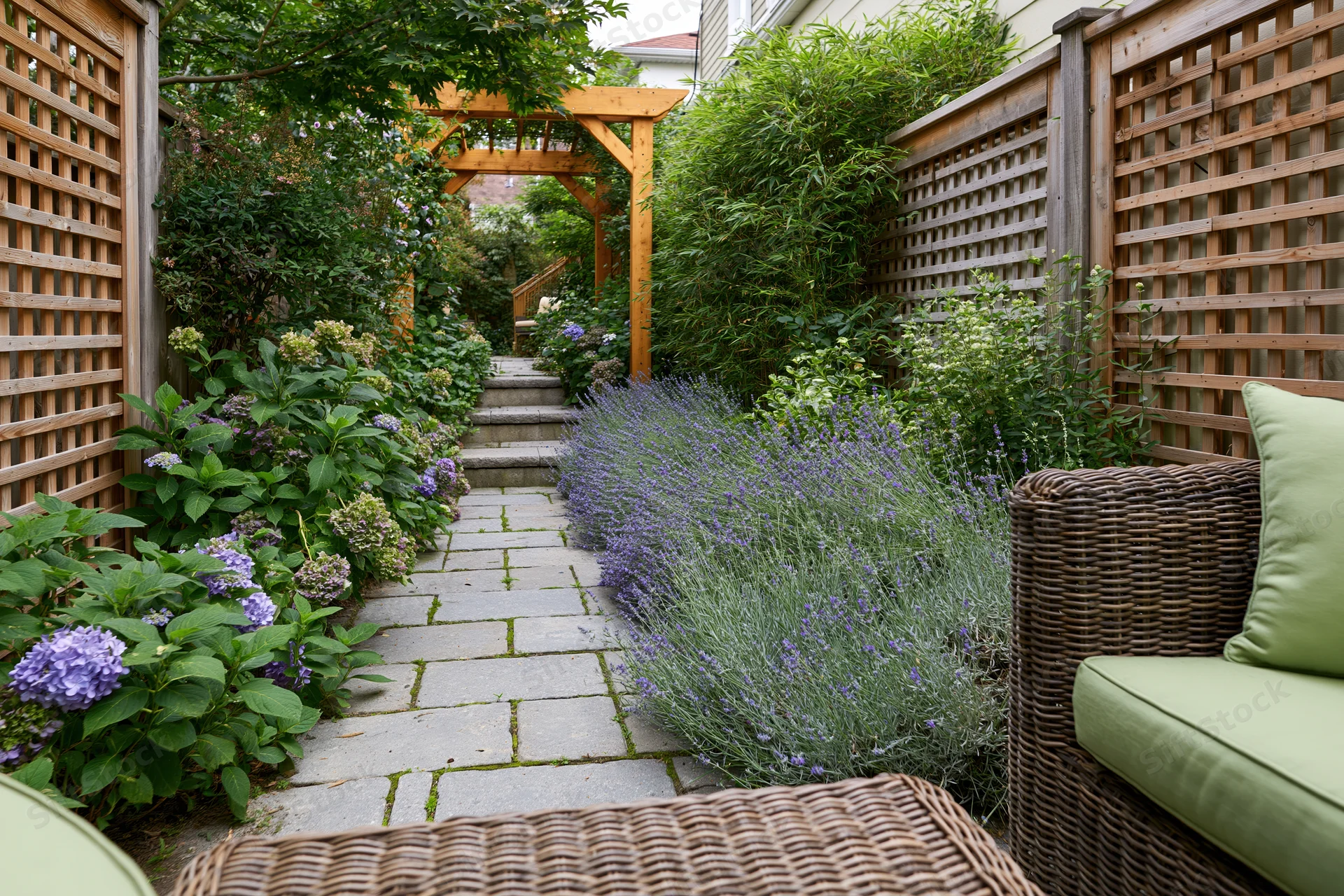Stone paver path lined with lavender and hydrangea in a narrow urban backyard garden with wicker furniture and wooden lattice fencing