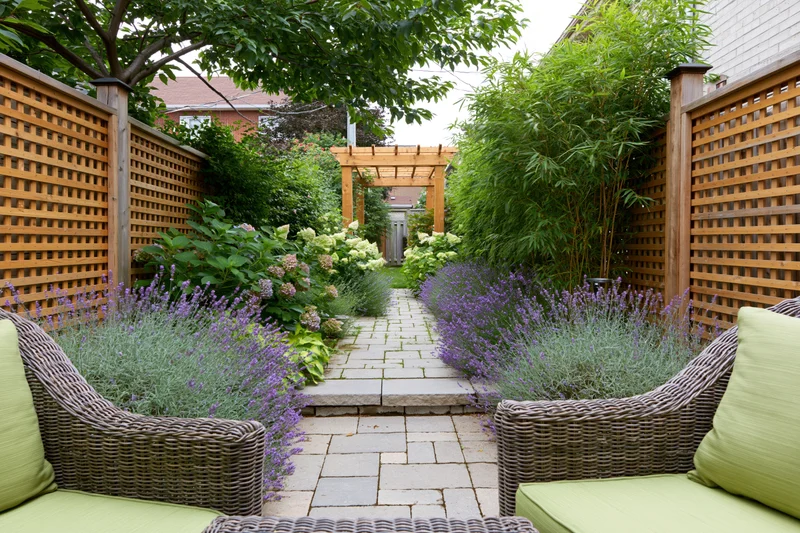 Narrow urban backyard garden with stone paver path leading through lavender and greenery to a wooden pergola, flanked by wicker chairs with green cushions and lattice fencing