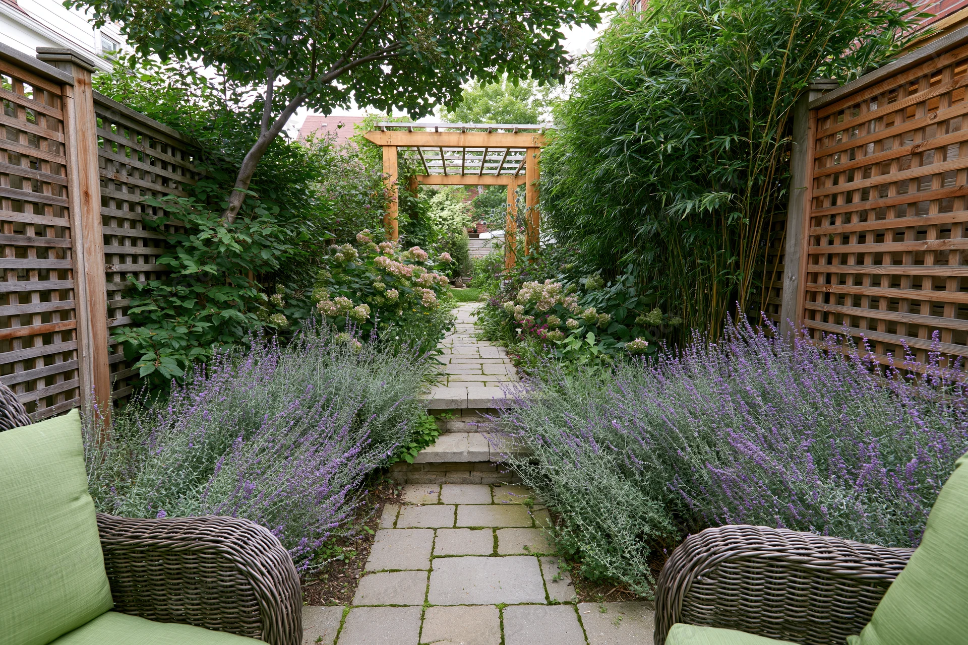 Narrow urban backyard garden with stone paver path lined with lavender and greenery, flanked by wicker seating and wooden lattice fencing, leading to a pergola in the distance
