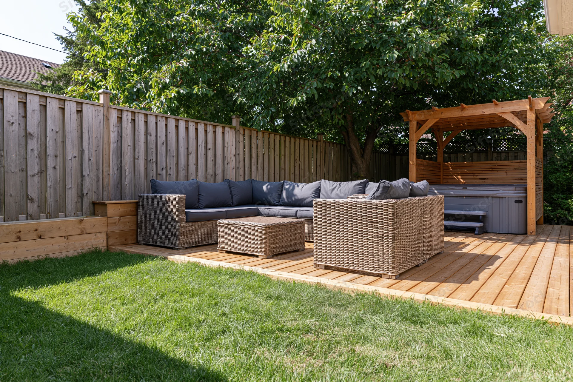 Wicker sectional sofa and coffee table on wooden deck in private backyard garden with hot tub under pergola and wooden fence