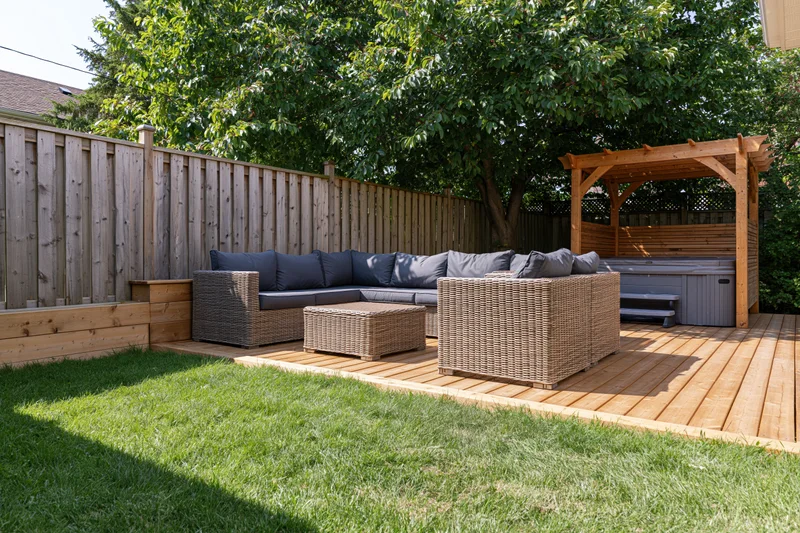 Wicker sectional sofa and coffee table on wooden deck in private backyard garden with hot tub under pergola and wooden fence