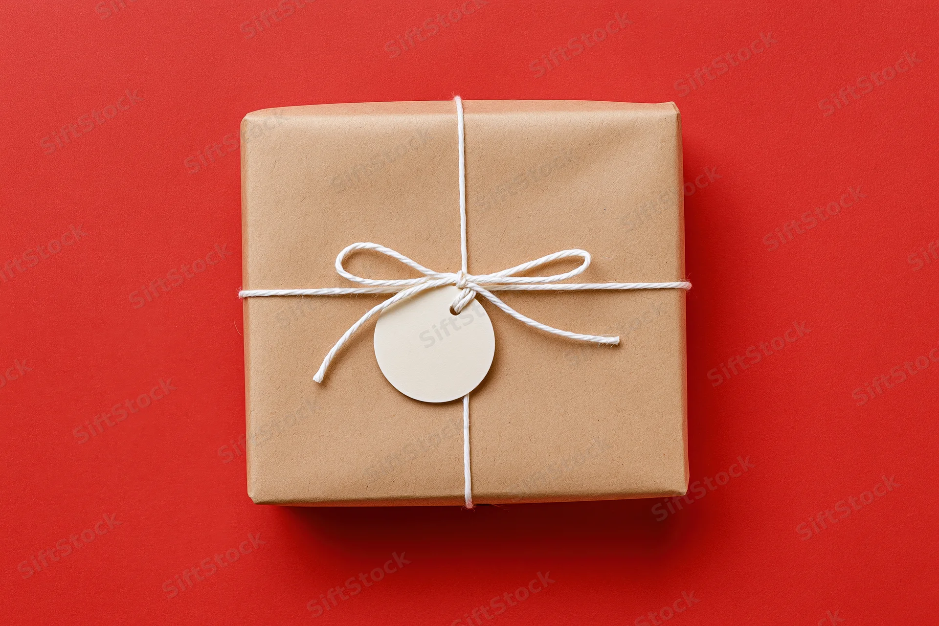 Rectangular gift box wrapped in brown kraft paper tied with white string and a blank circular tag on red background