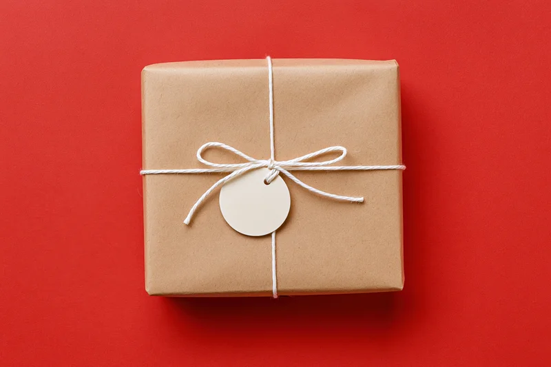 Rectangular gift box wrapped in brown kraft paper tied with white string and a blank circular tag on red background