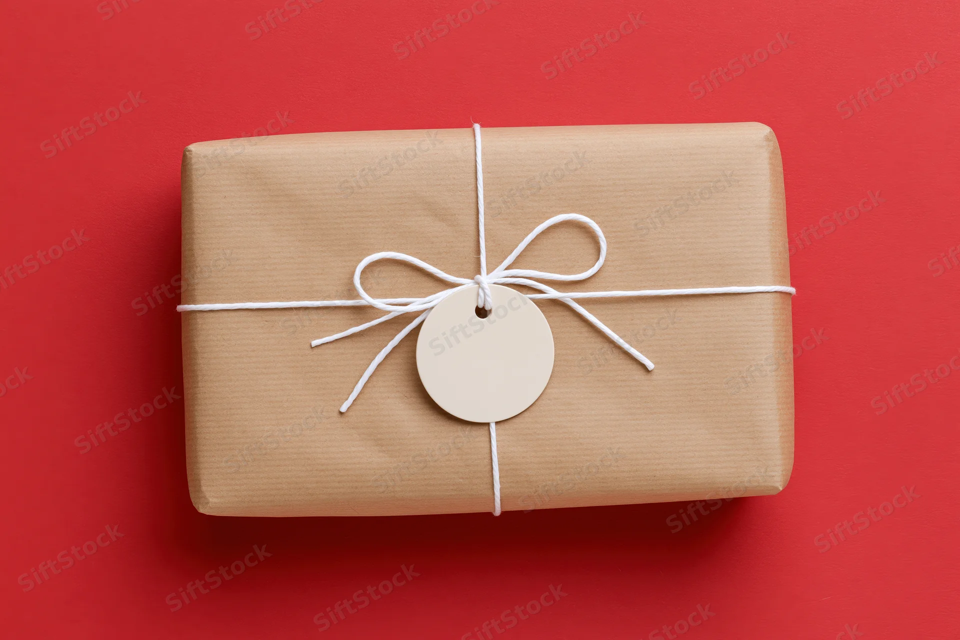 Rectangular gift box wrapped in brown kraft paper tied with white string and a blank circular tag on red background