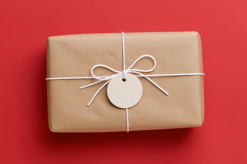Rectangular gift box wrapped in brown kraft paper tied with white string and a blank circular tag on red background