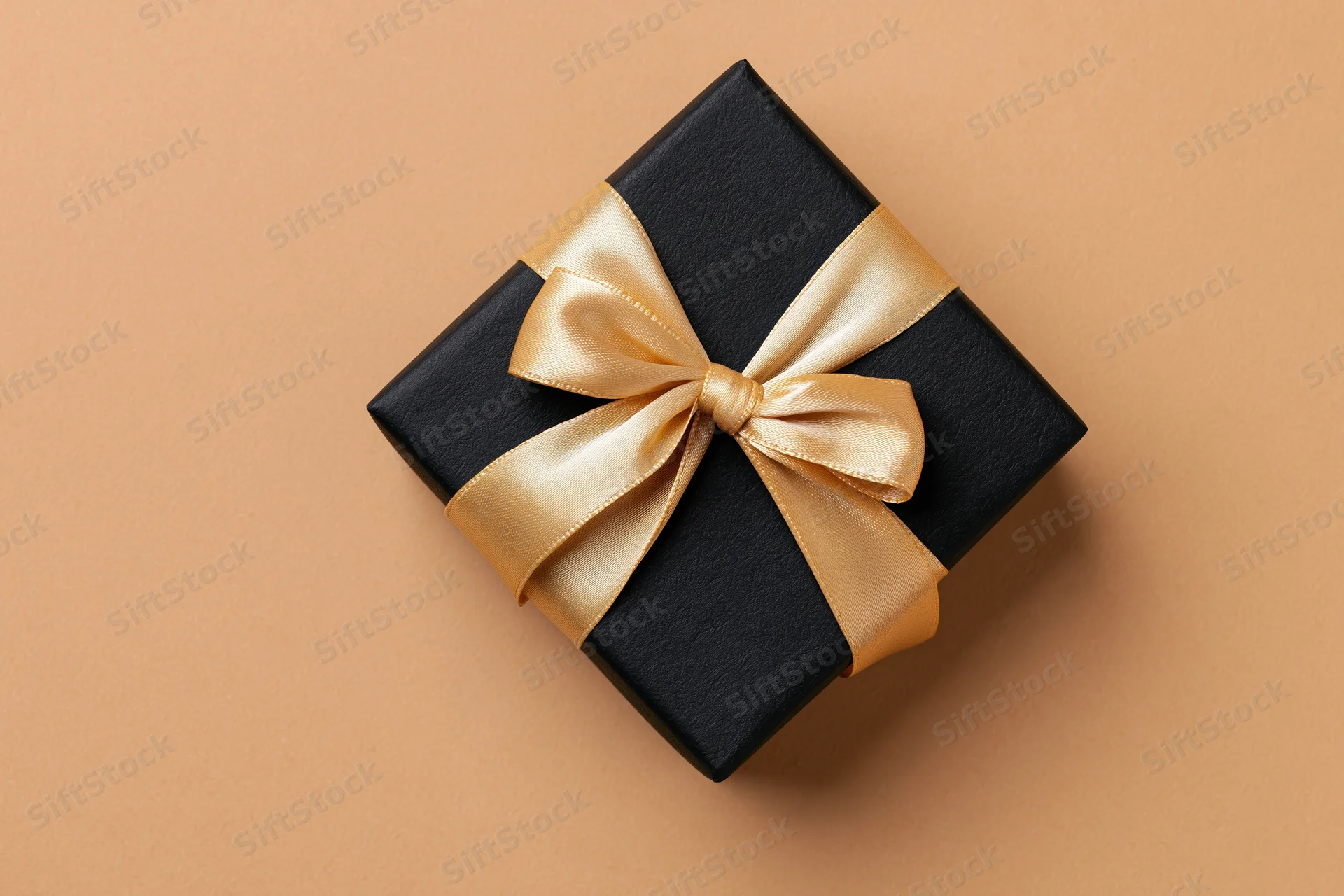 Black gift box wrapped with gold satin ribbon on beige surface