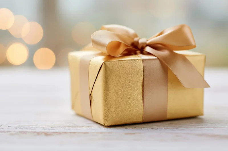 Gold-wrapped gift box with ribbon bow on wooden table, soft bokeh lights in background