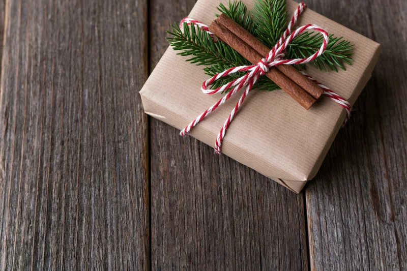 Small gift box wrapped in kraft paper with red and white twine, pine sprig and cinnamon sticks on wooden surface