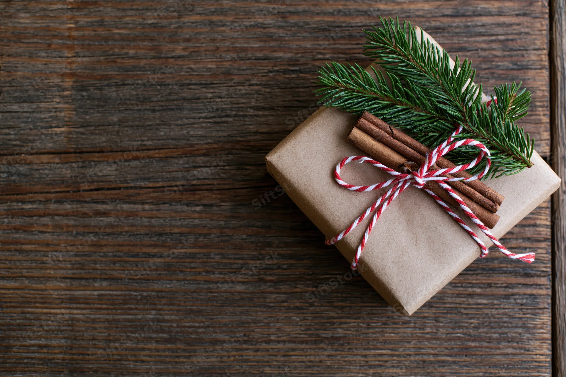 Small gift wrapped in kraft paper with red and white twine, cinnamon sticks and pine branch on wooden surface