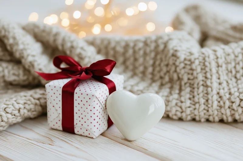 Small gift box with red ribbon and white heart ornament on wooden table beside knit blanket with bokeh lights in background