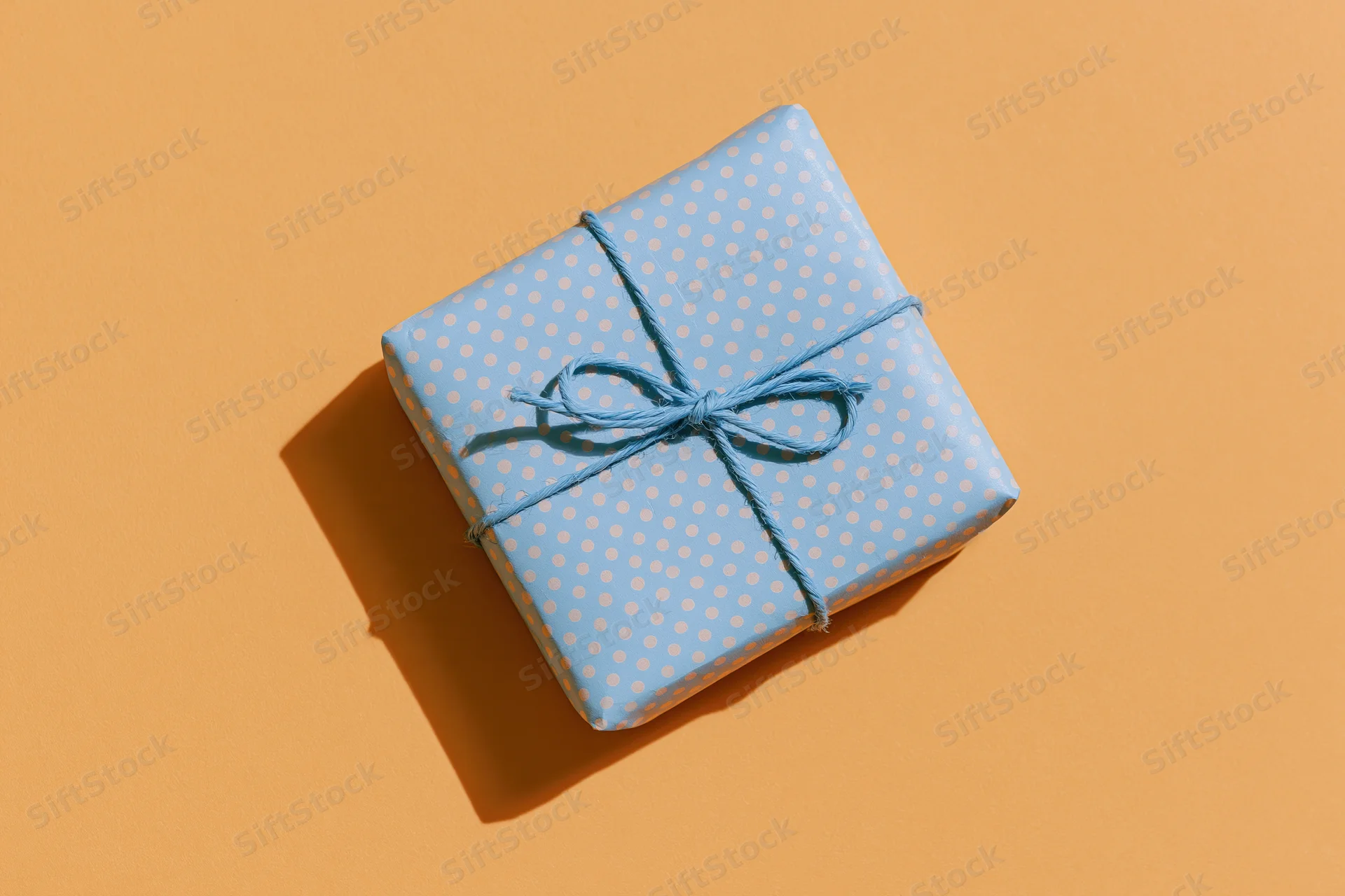 Small gift box wrapped in blue polka dot paper with string bow on orange background