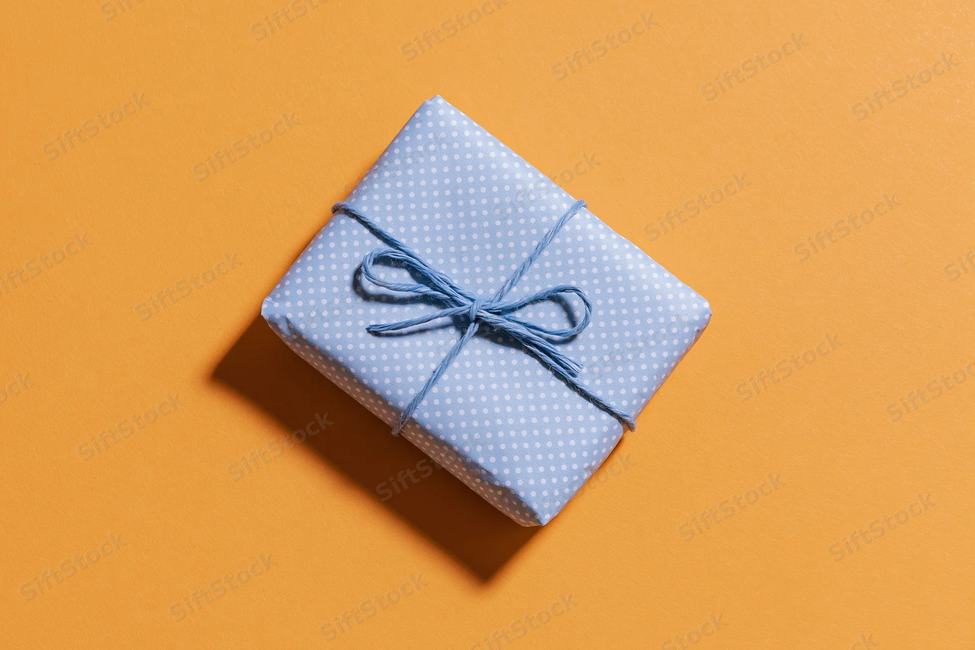 Small gift box wrapped in blue polka dot paper with string bow on orange background