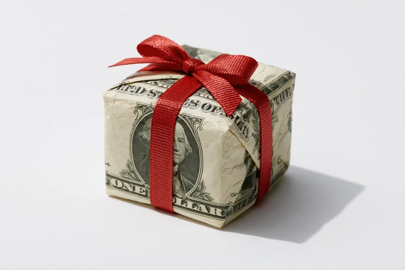 Small cube-shaped gift wrapped in us dollar bills with red ribbon bow on white background