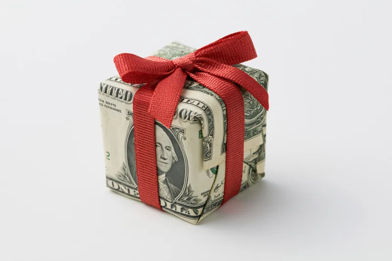Small gift box wrapped in us dollar bills with red ribbon on white background