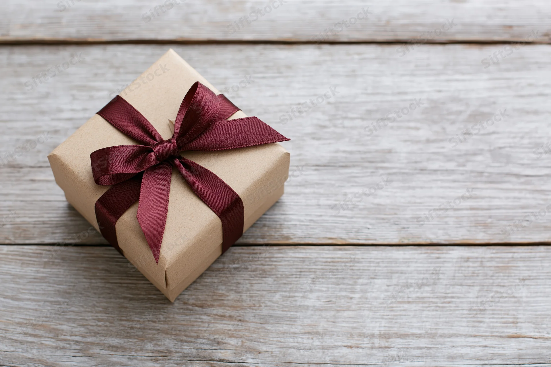 Small gift box wrapped in kraft paper with burgundy ribbon bow on rustic wooden surface