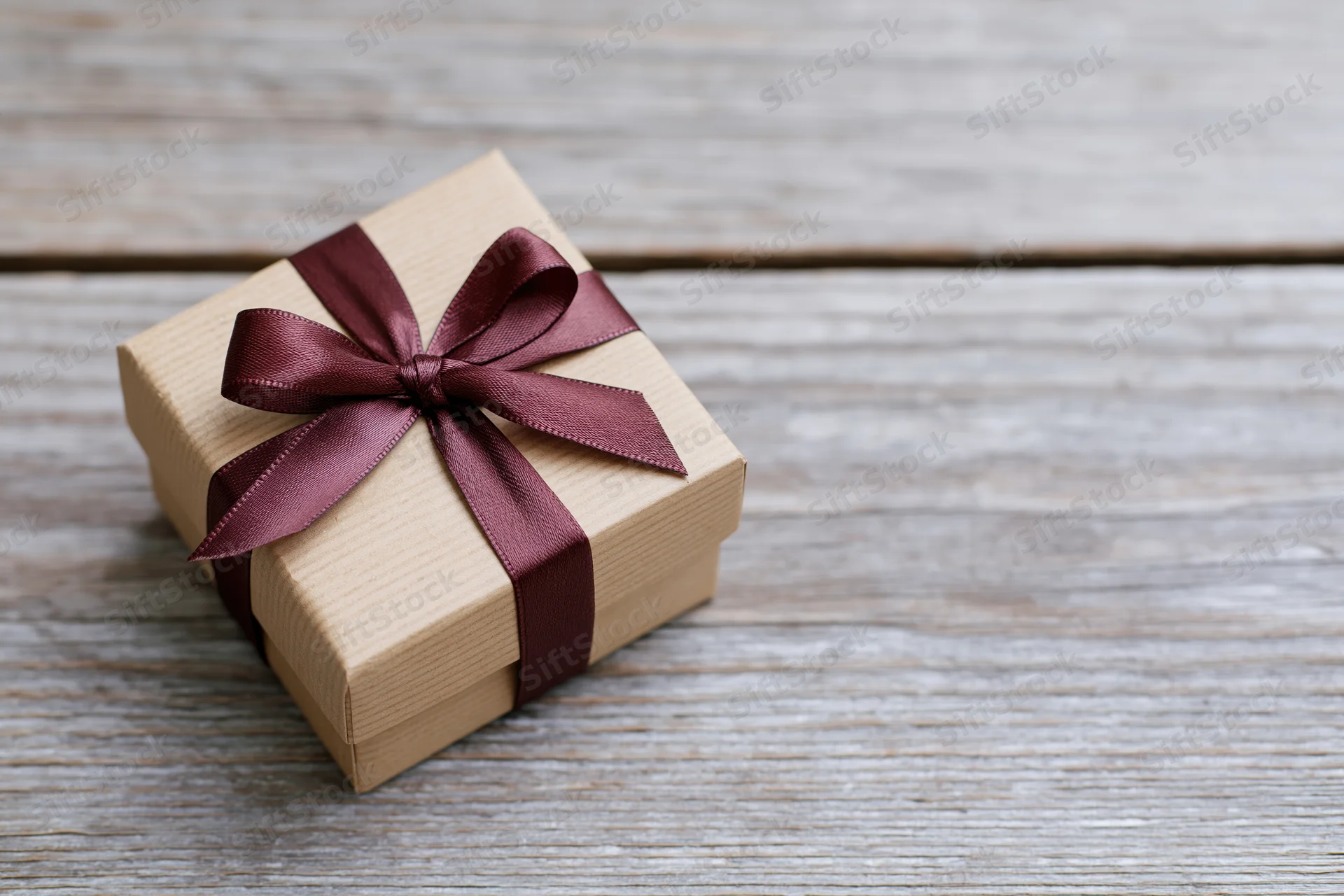 Small gift box wrapped in kraft paper with burgundy ribbon bow on wooden surface