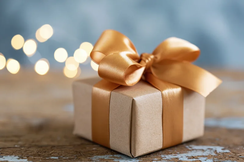 Small gift box wrapped in kraft paper with golden ribbon bow on wooden surface, blurred lights in background