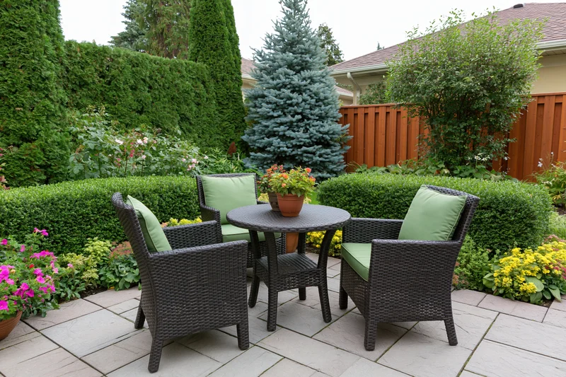 Dark wicker chairs and table with green cushions arranged on a stone patio surrounded by shrubs and flowering plants in a suburban backyard