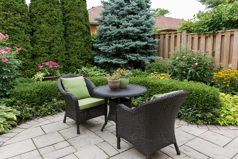 Dark wicker chairs and table on a stone patio surrounded by lush greenery and flowering plants in a suburban backyard
