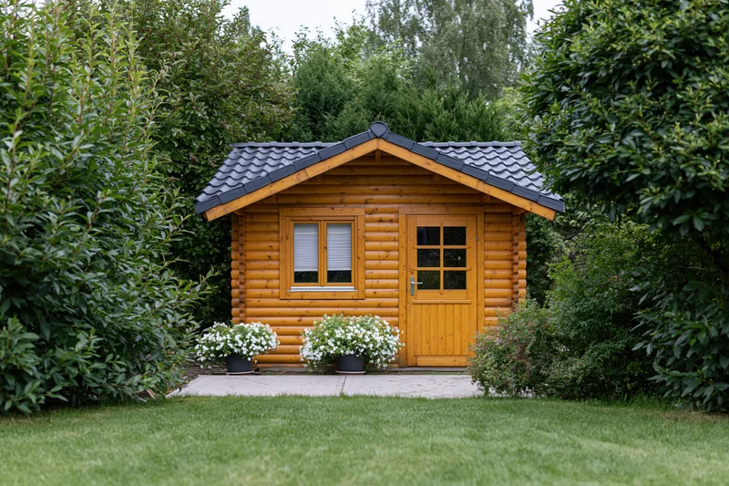 Small wooden garden cabin with tiled roof surrounded by green shrubs and white flowers on a lawn
