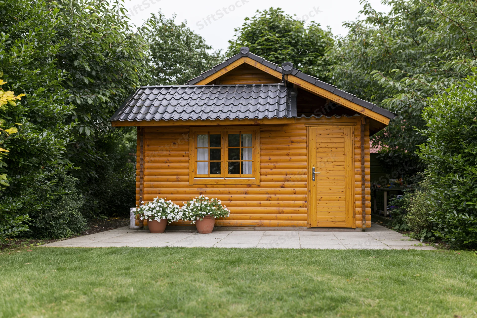 Small wooden garden cabin with tiled roof and potted flowers on patio surrounded by greenery