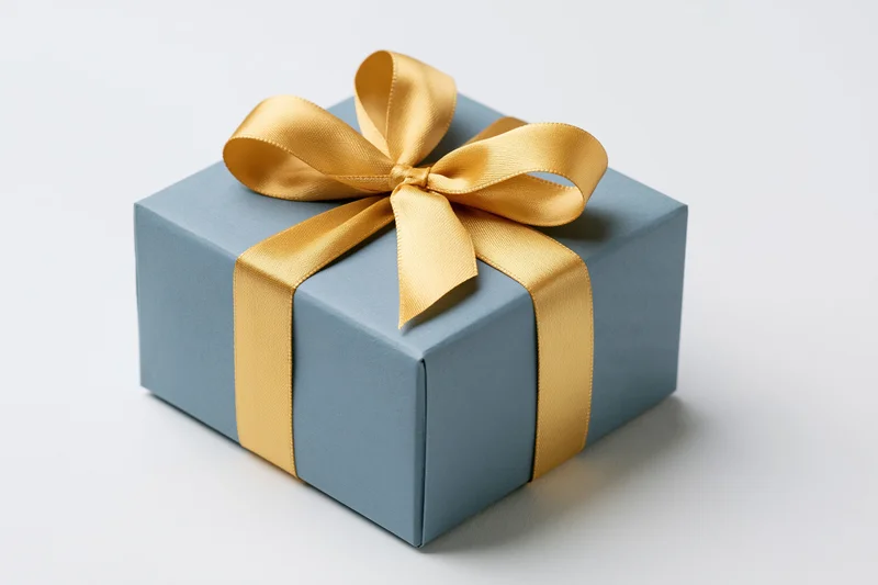 Square gift box wrapped in blue paper with gold ribbon bow on white background