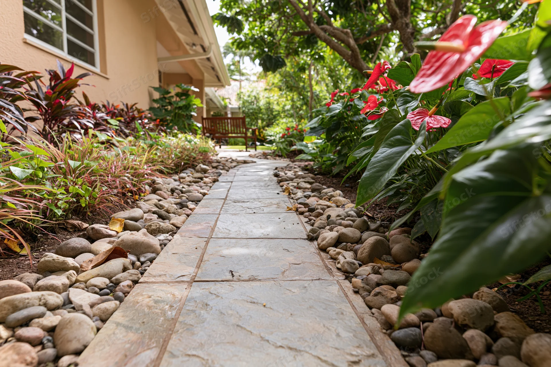 Stone tile garden path lined with river rocks and tropical plants leading to a wooden bench in a shaded backyard
