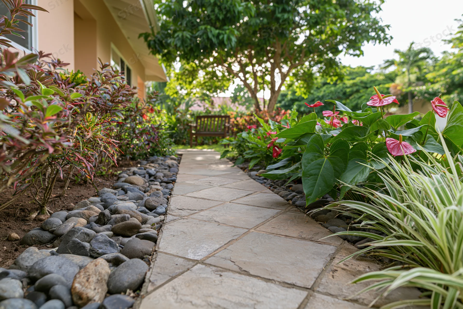 Stone tile garden path lined with river rocks and flowering plants leading to a wooden bench near a house