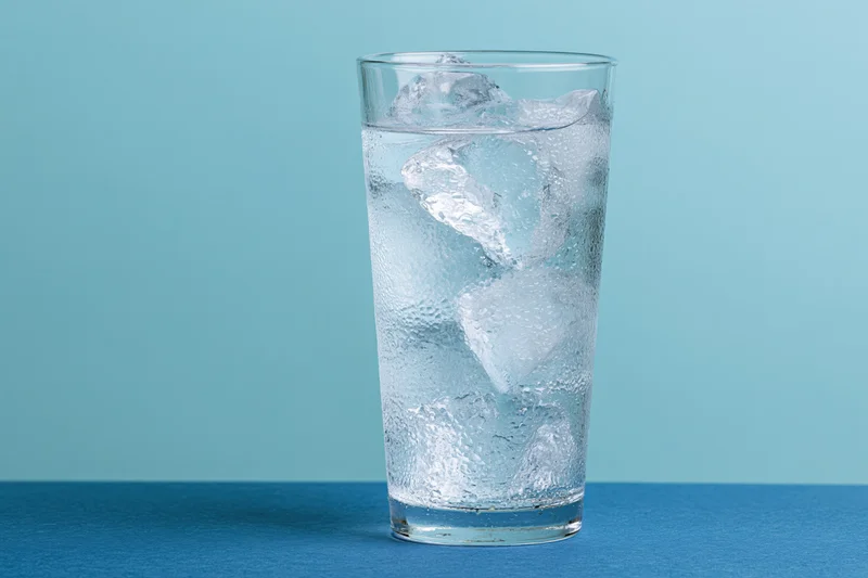 Tall glass filled with ice water and large ice cubes on blue surface