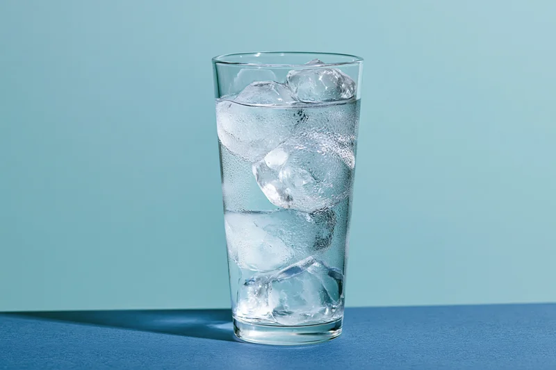 Tall glass filled with ice water and large ice cubes on blue surface