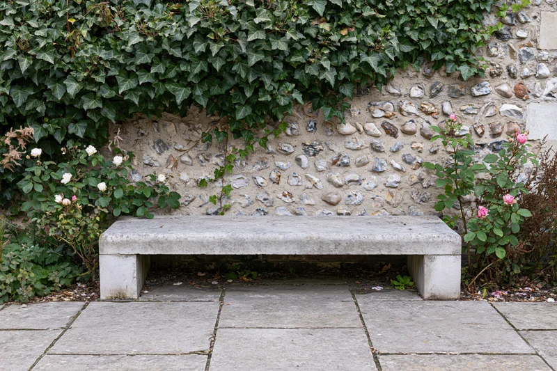 Weathered stone bench sits empty against ivy-covered stone wall in garden courtyard