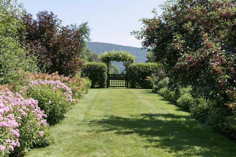 Mown grass pathway lined with flowering shrubs and hedges leading to a gated arbor in a private garden