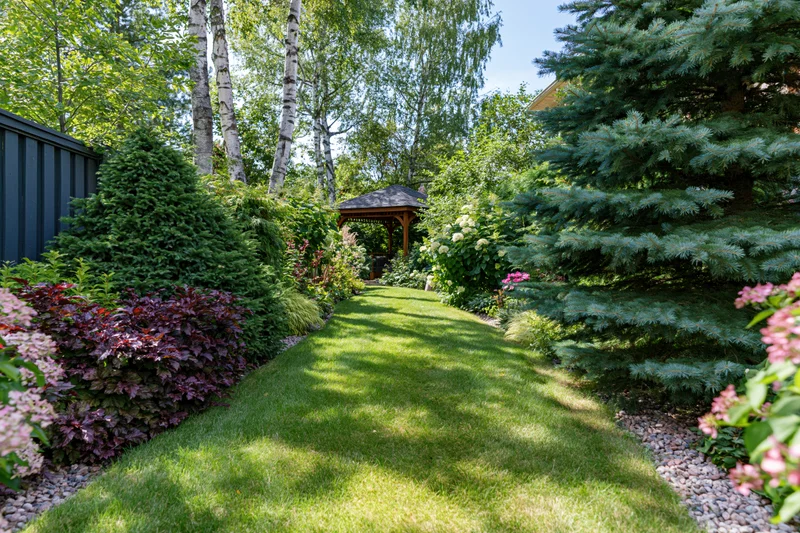 Narrow green lawn pathway through lush garden bordered by trees and flowering shrubs leading to wooden gazebo